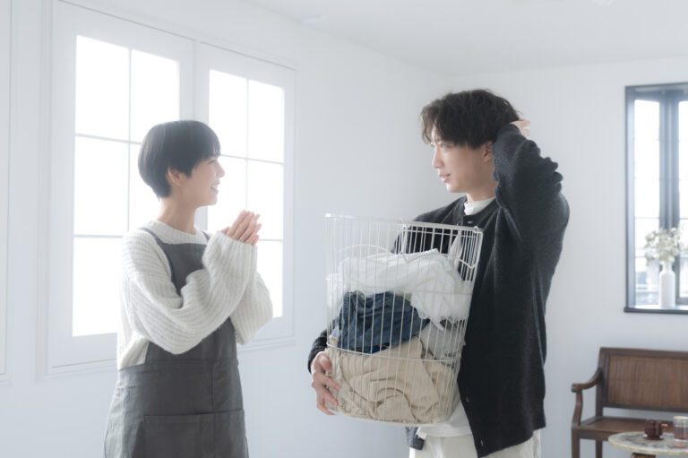 A woman in an apron clasps her hands toward a man holding a full laundry basket in a bright room