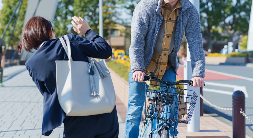 Woman with a white tote bag flinching and raising her hand as a man on a bicycle passes close to her on a sidewalk