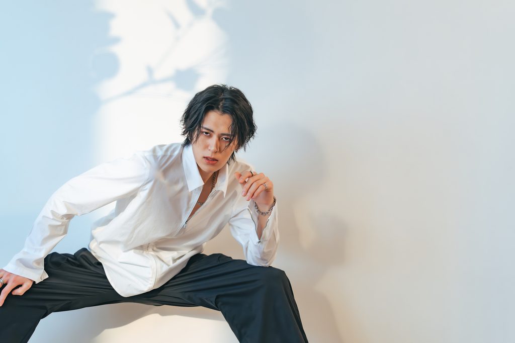 Young Japanese man in a loose white shirt and black trousers crouching against a pale wall, staring at the camera