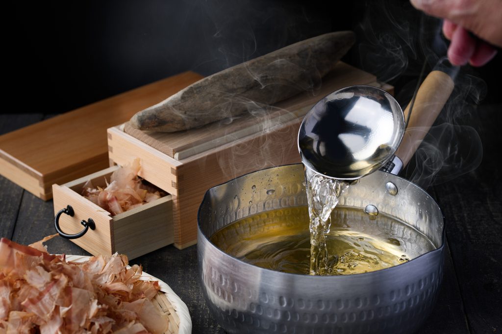 A ladle pouring dashi into a pot beside shaved bonito flakes and a block of katsuobushi on a wooden shaver box.