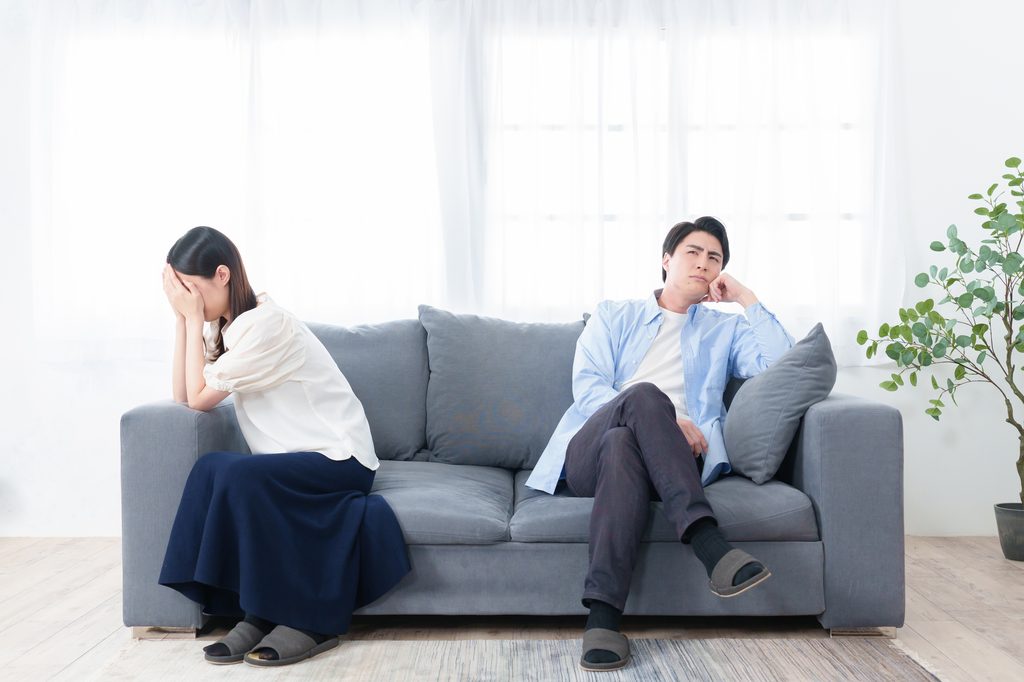 A woman covers her face with her hands at one end of a sofa while a man sits at the other end looking away