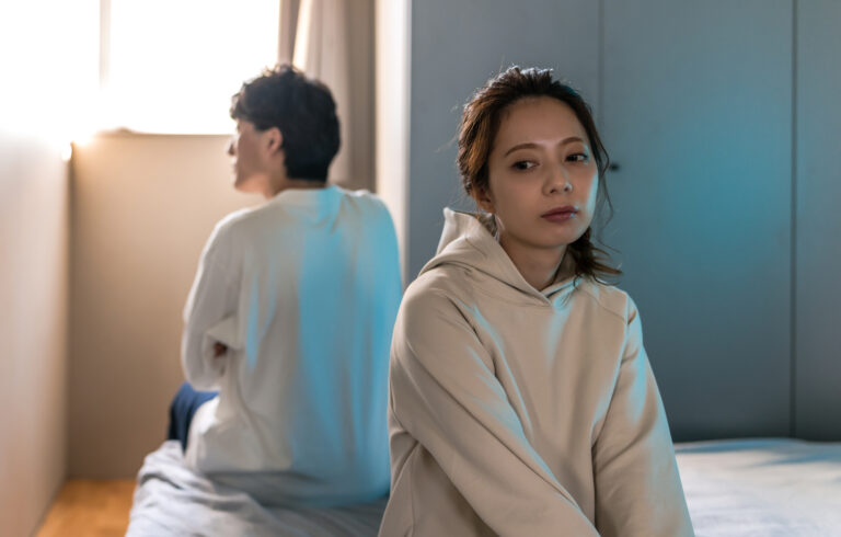 A woman sits on the edge of a bed looking away, while a man sits behind her with his back turned