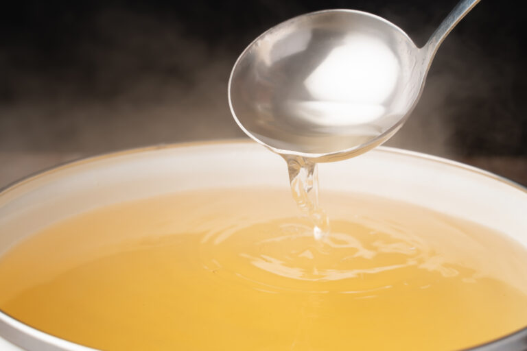 A ladle pouring clear golden broth into a steaming pot of stock.
